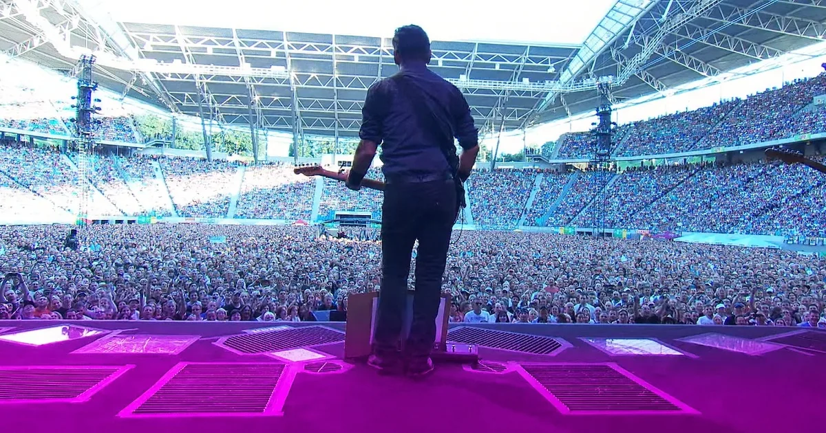A musician stands on stage with a guitar, facing a large, enthusiastic crowd in a stadium.
