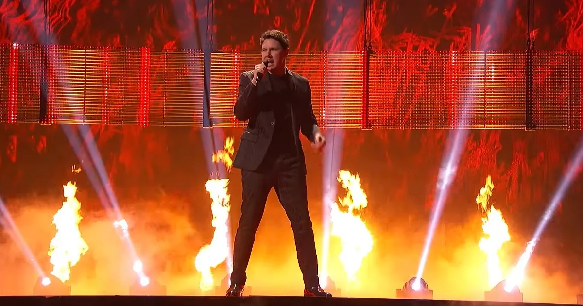 A man in a black suit sings on stage with red lights and fire effects.
