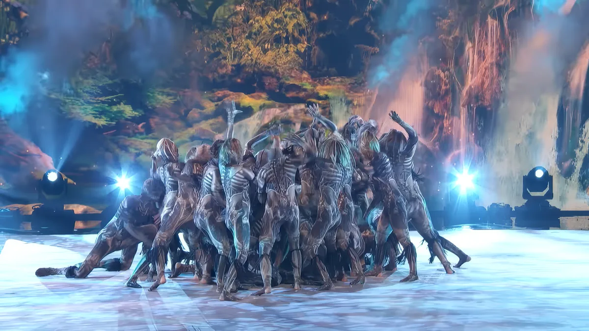 A group of performers in metallic costumes are huddled together on stage, surrounded by dramatic lighting and a forest...