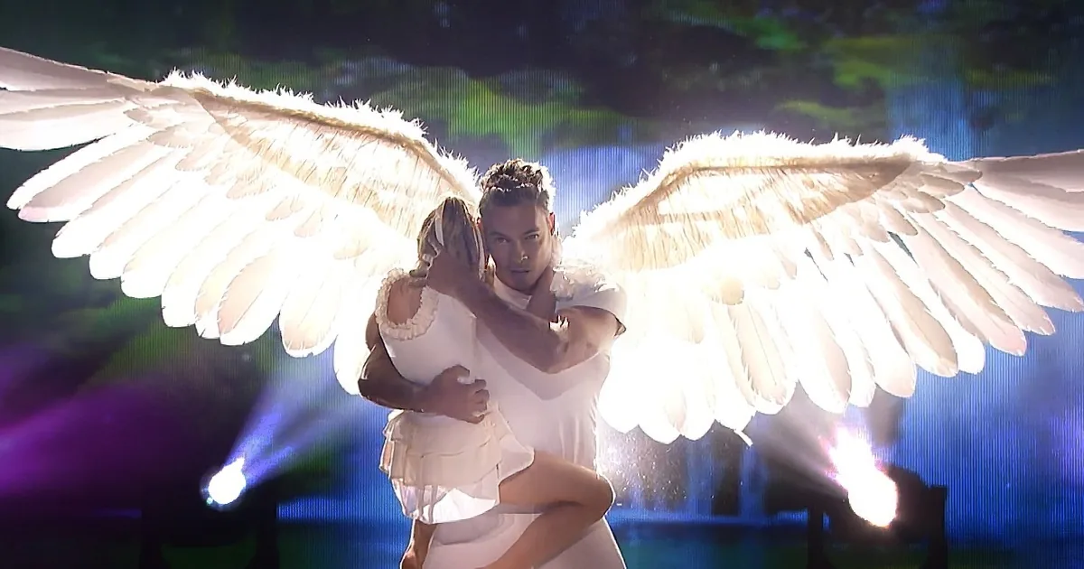 A man and woman in white, with angel wings, embrace on stage under bright lights.