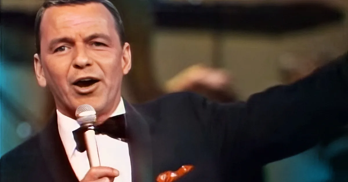A man in a suit and bow tie sings into a microphone, gesturing with his arm.
