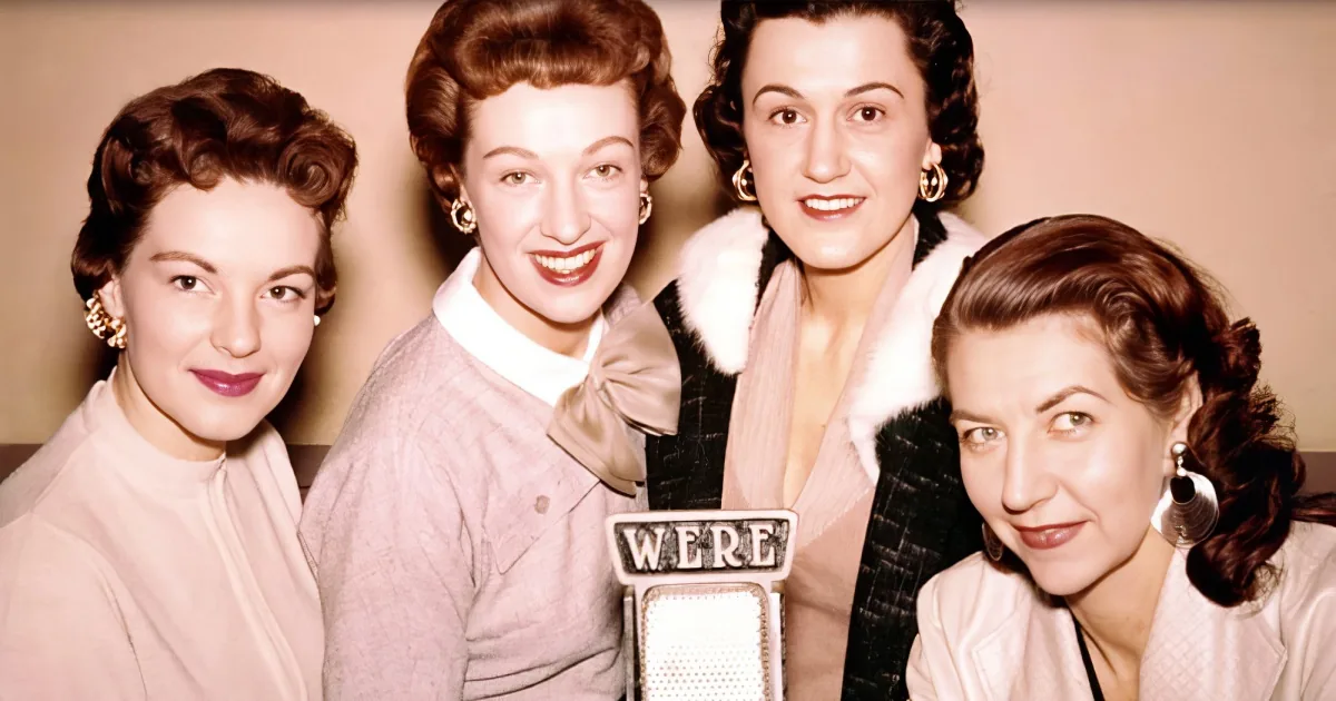 Four women in vintage attire pose together, smiling, with one holding a microphone.