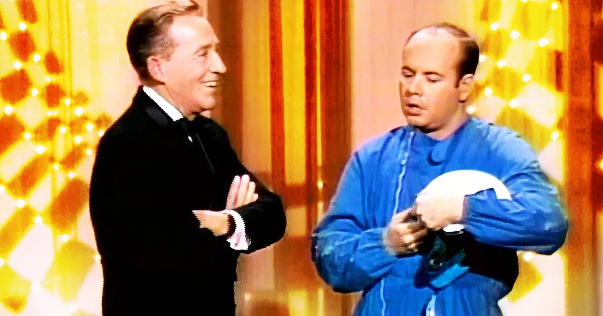 A man in a tuxedo smiles while another in a blue outfit adjusts his tie, both against a golden backdrop.