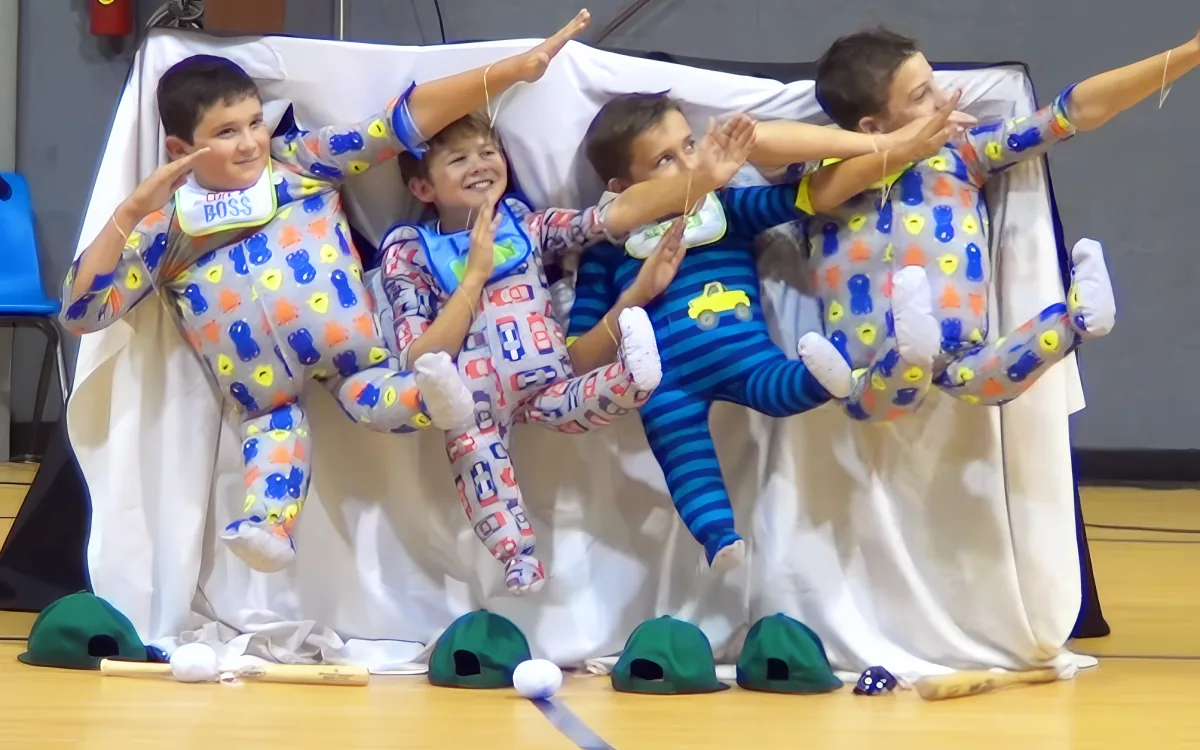 Four children in colorful pajamas perform a synchronized dance on a gym floor with hats and a baton.