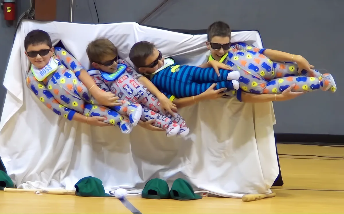 Four children in pajamas are being held up by a person in a white cloth, wearing sunglasses and holding a stuffed animal.
