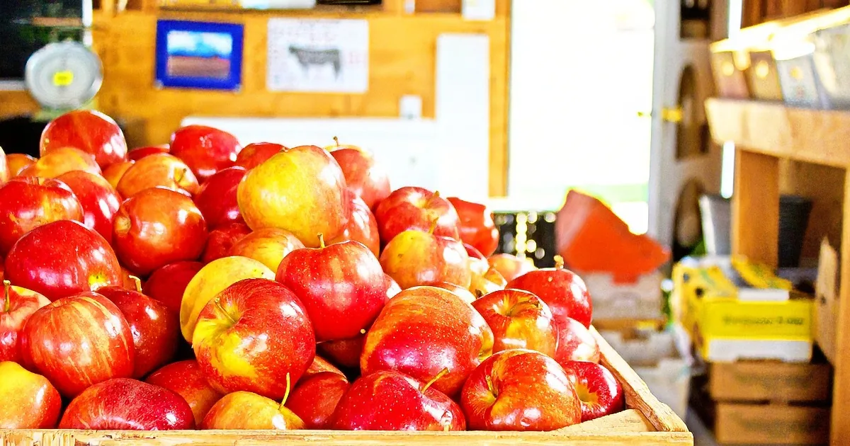 A pile of red apples in a wooden crate, with a blurred background of a store interior.