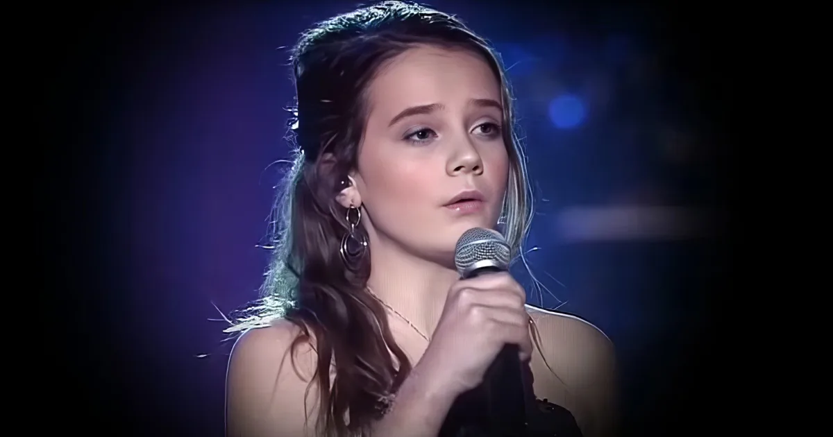 A woman with long hair and large hoop earrings sings into a microphone on stage.