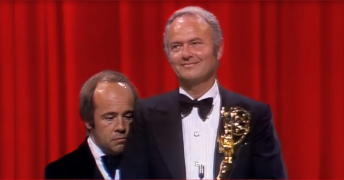 A man in a tuxedo holding an Oscar statue, smiling, with a red curtain backdrop.