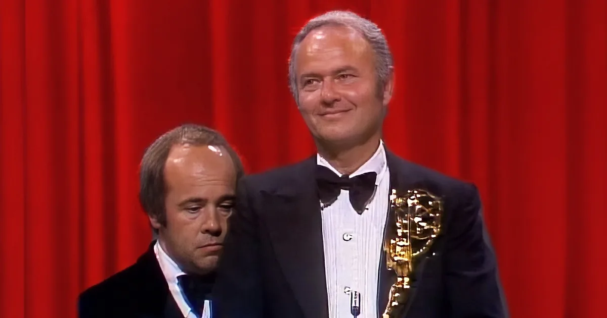 A man in a tuxedo holds an Emmy award, standing next to another man in a tuxedo, both against a red curtain backdrop.