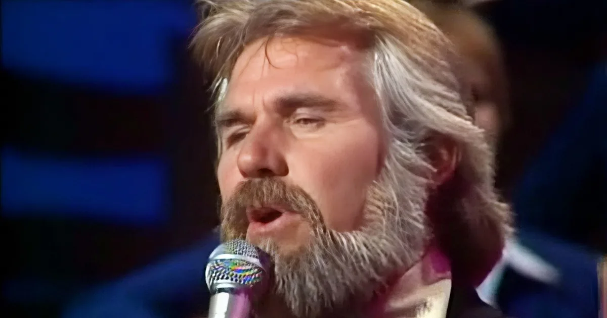A man with long, gray hair and a beard sings into a microphone, wearing a suit.