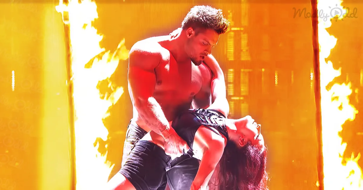 A muscular man in jeans holds a woman upside down, both in a dramatic, fiery setting.