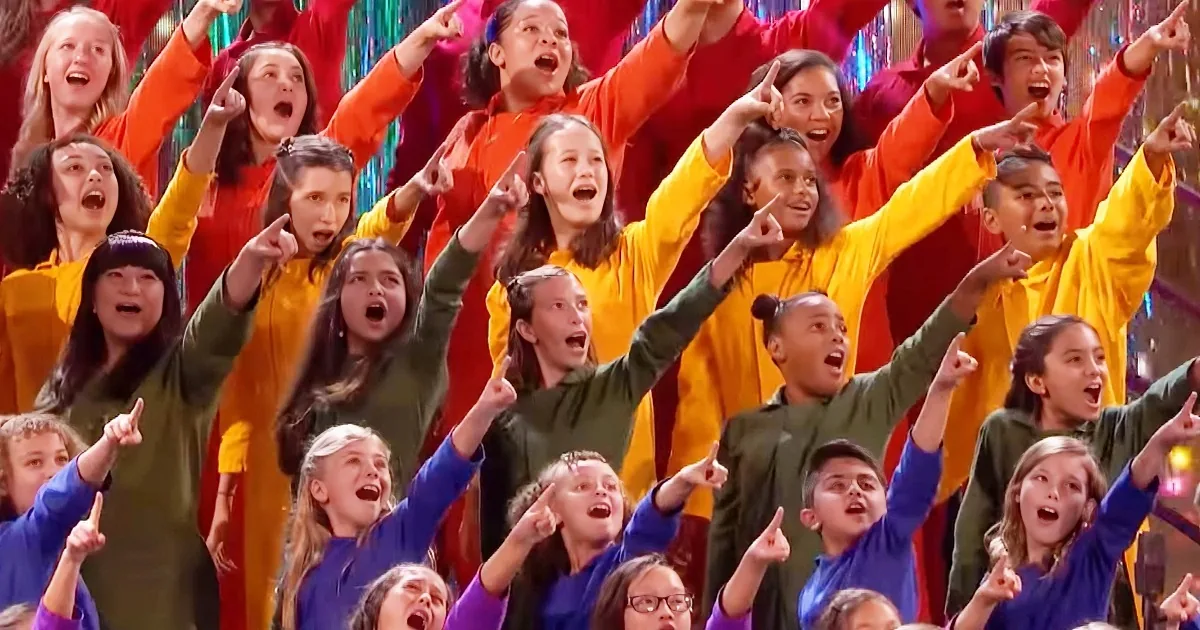 A group of children in colorful outfits are enthusiastically raising their hands and pointing upwards.