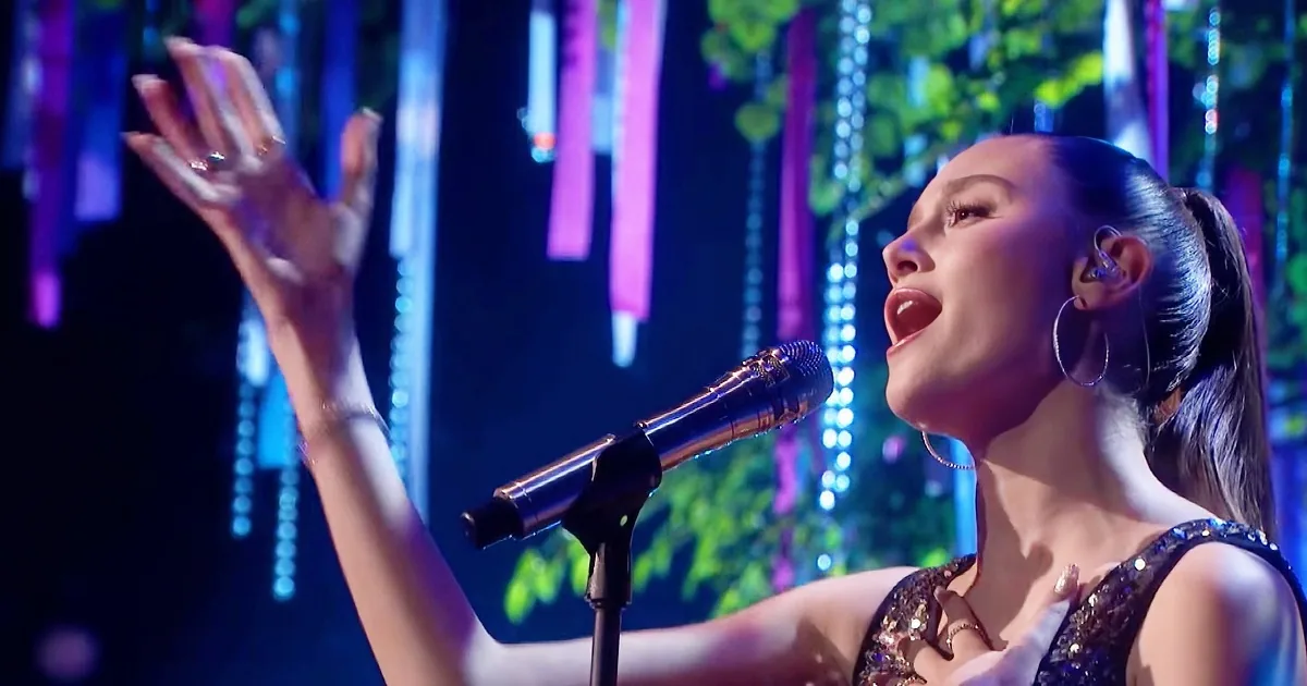 A singer with a microphone, raising her hand, wearing a sparkly outfit, with colorful lights in the background.