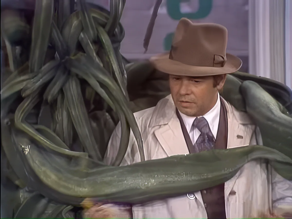 A man in a fedora and trench coat stands beside a large, green, snake-like creature.