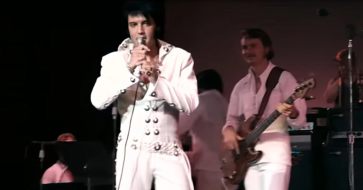 Elvis Presley in a white suit sings into a microphone on stage.