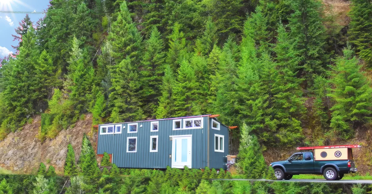 A blue tiny house on wheels is parked on a road surrounded by lush green trees.