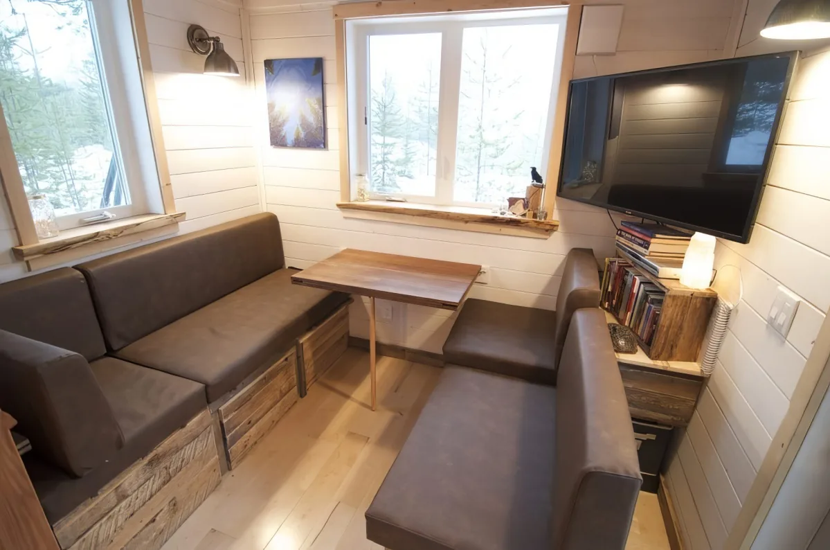 A cozy, rustic living space with a sofa, small table, books, and a TV, all set against a backdrop of snowy trees.