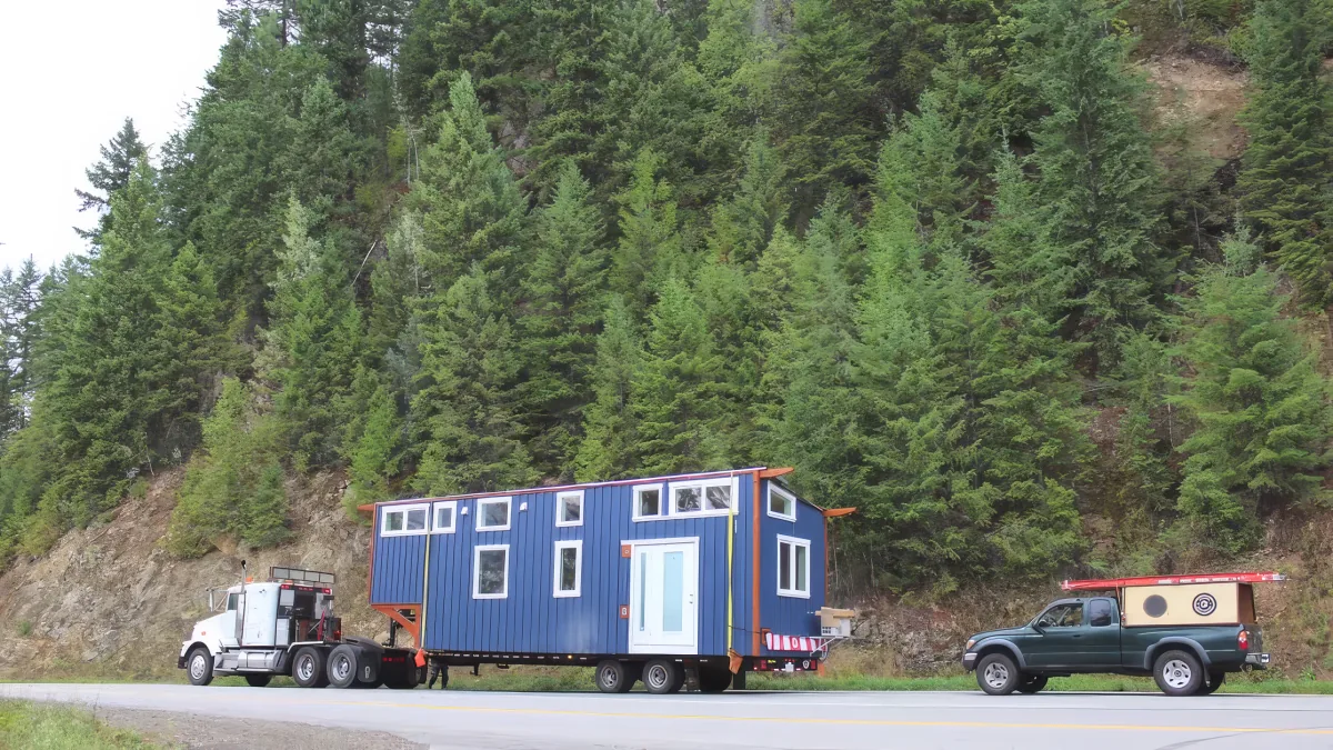 A large blue modular home is being transported on a flatbed truck by a semi-truck on a road surrounded by dense green trees.