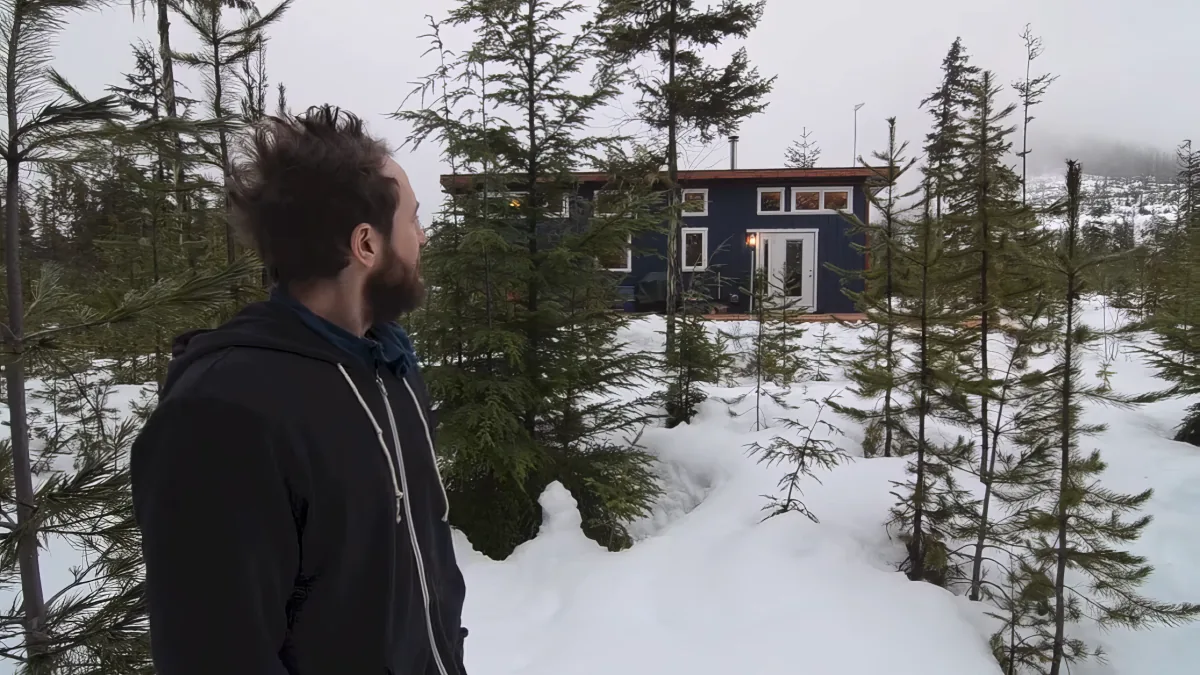 A man with a beard stands in a snowy forest, gazing at a blue cabin with a red roof.