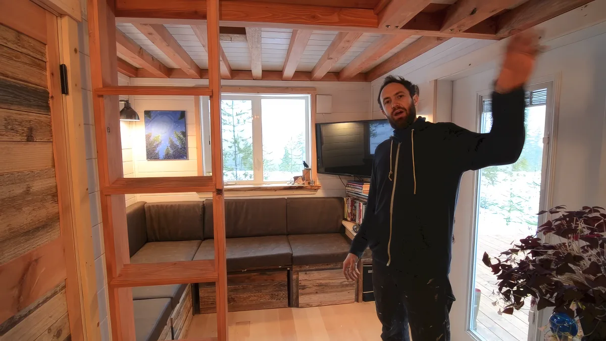 A man in a black hoodie gestures upwards in a cozy, rustic room with wooden beams, a couch, and large windows.