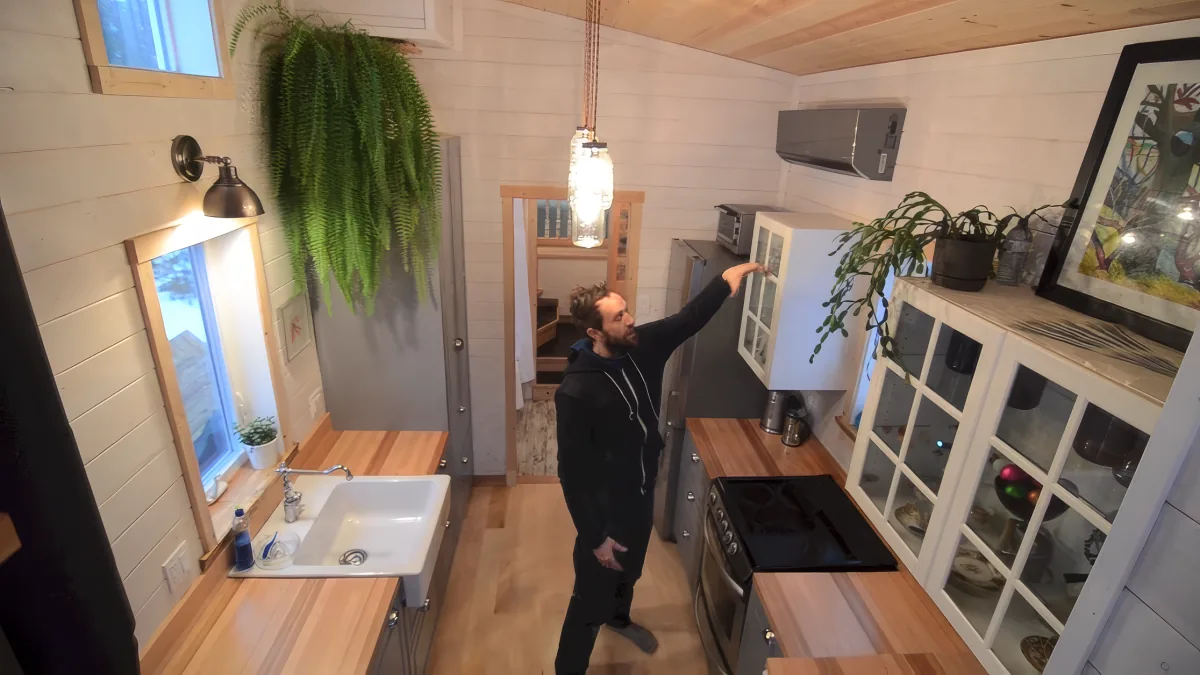 A man stands in a compact, well-lit kitchen with wooden cabinets, a sink, and a hanging plant.