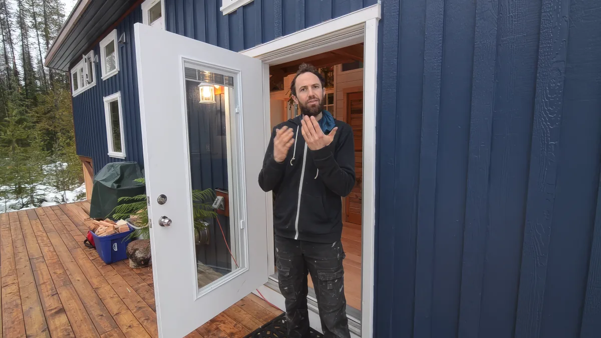 A man stands in front of a blue, wooden cabin, gesturing with his hands as he speaks.