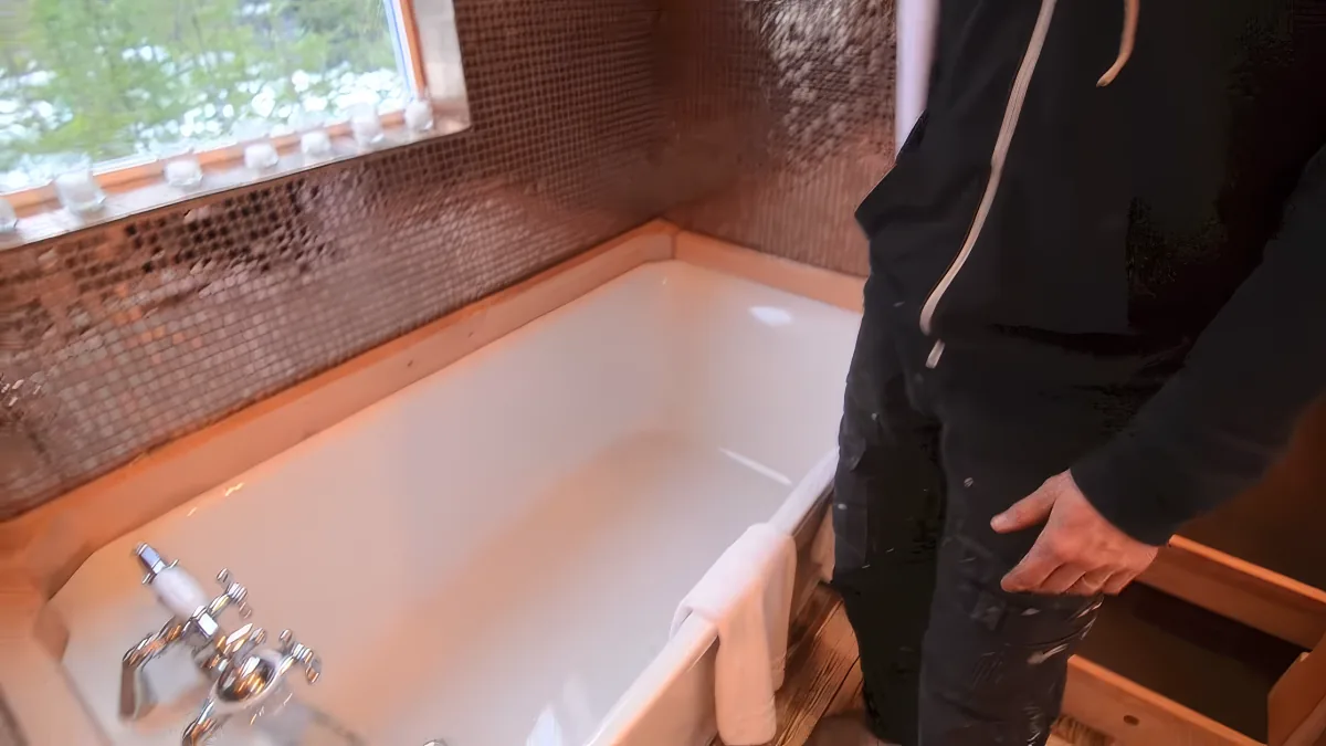 A person in black pants and a hoodie stands beside a white bathtub with a mosaic tile backsplash and a window above.