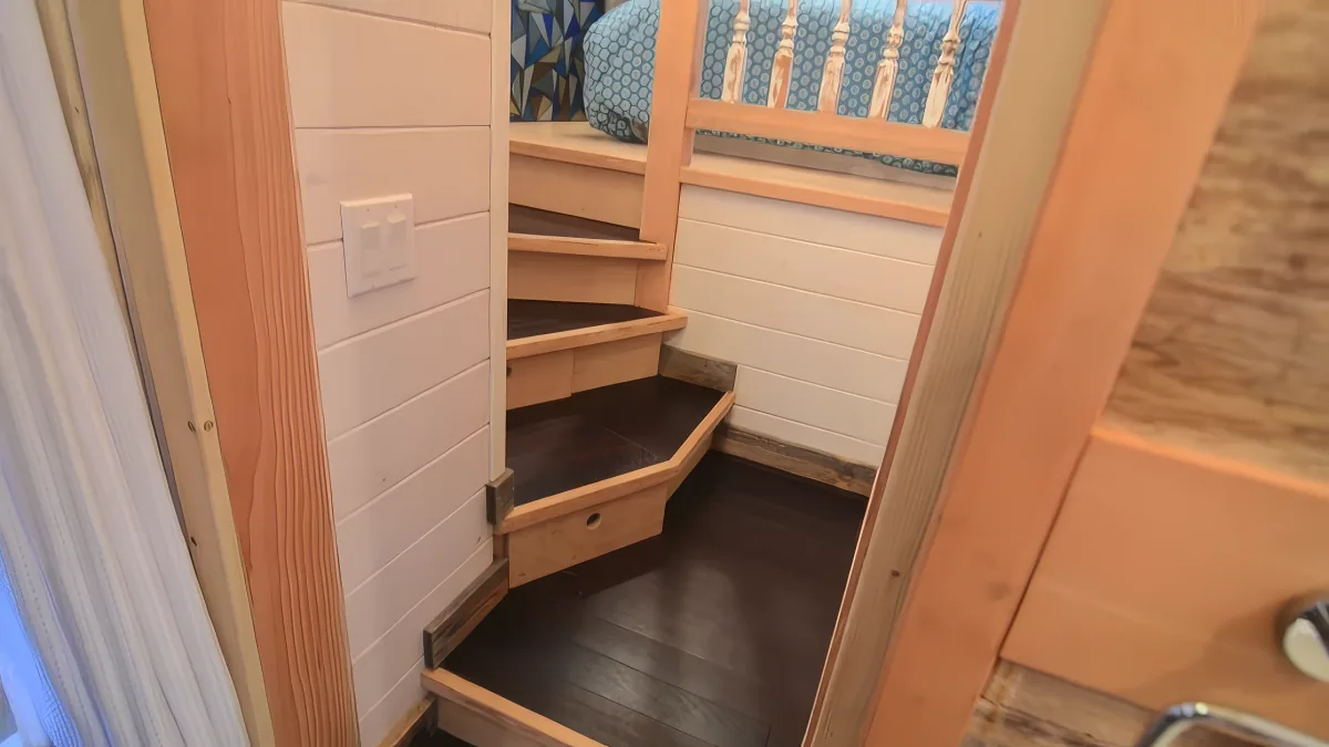 A wooden staircase leads up to a loft bed with a blue patterned blanket.