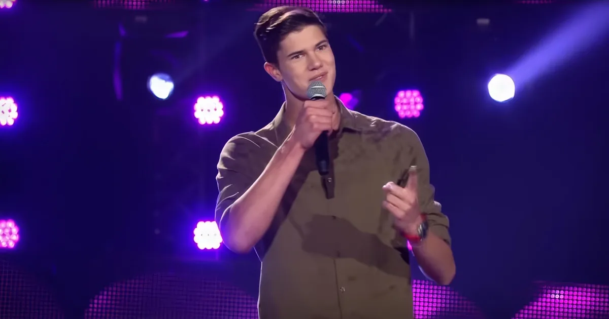 A person in a brown shirt holds a microphone on stage with purple lights in the background.