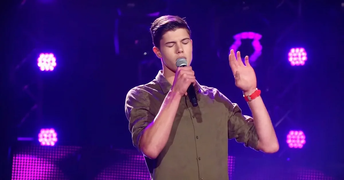 A young man sings into a microphone on stage with purple lights behind him.