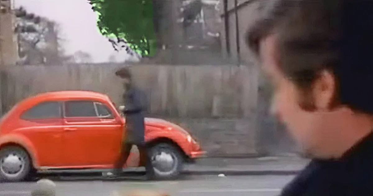 A man in a black shirt observes a person in a blue shirt standing next to a bright orange Volkswagen Beetle.
