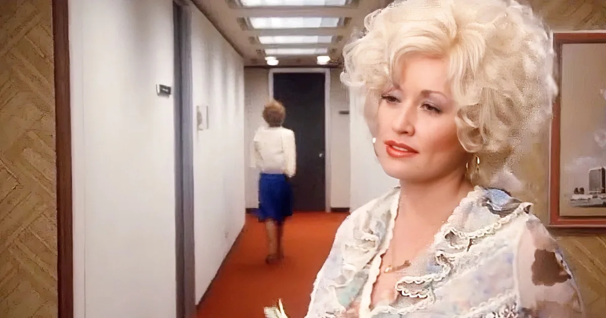 A woman with blonde, curly hair and a floral blouse walks down a hallway, looking back.