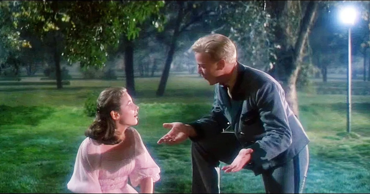A man in a dark jacket gestures towards a woman in a pink dress in a grassy park at night.