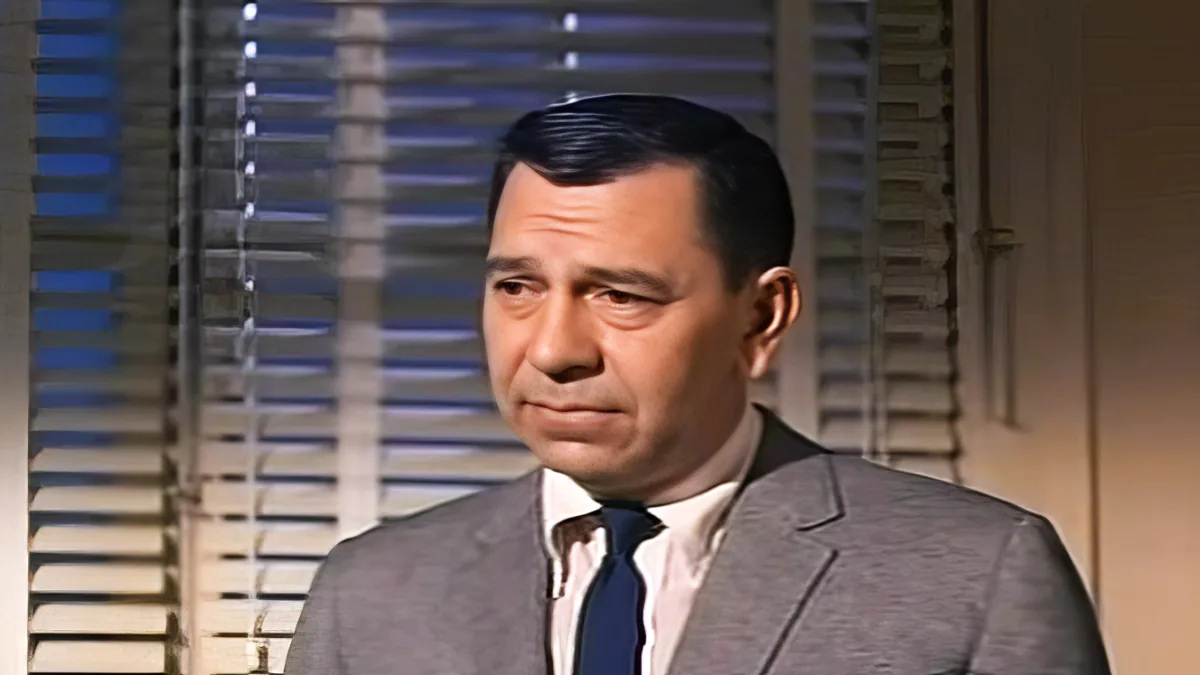 A man in a suit and tie is speaking, with blinds in the background.