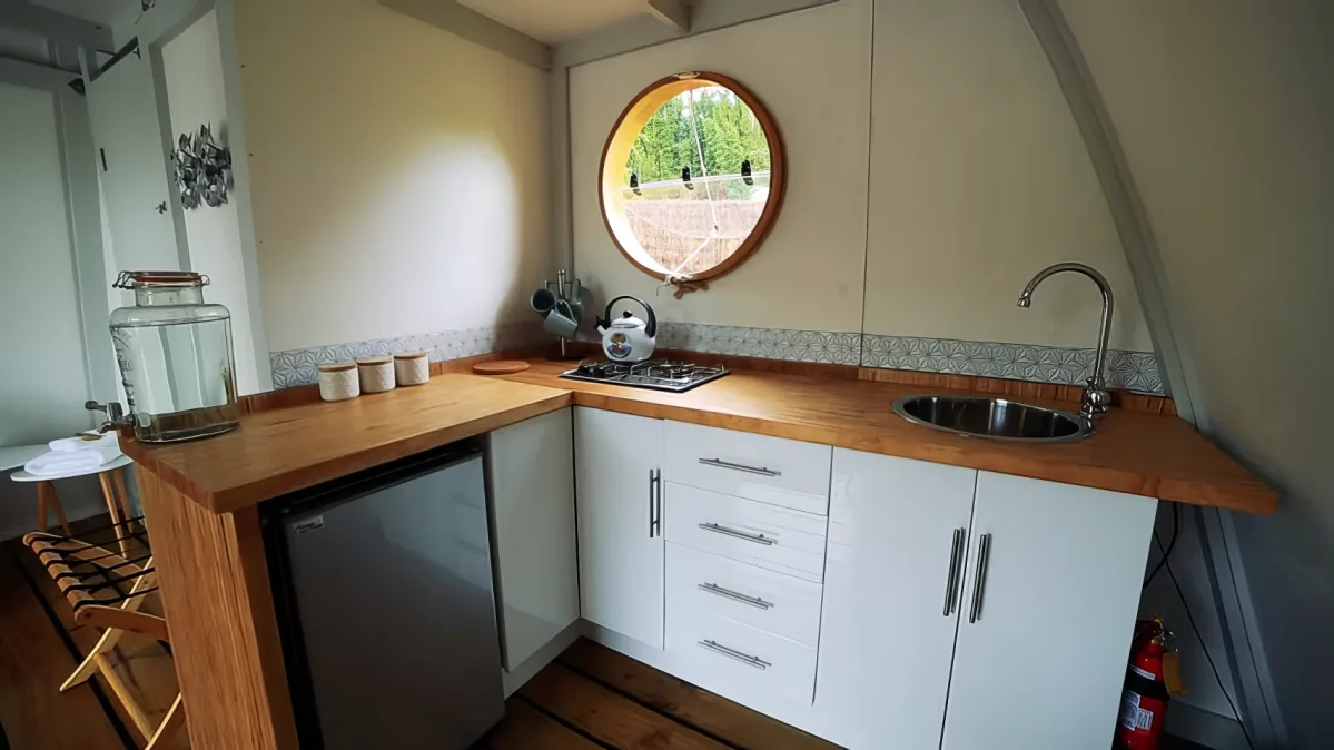 A compact kitchen with white cabinets, wooden countertop, sink, and round window showing greenery outside.