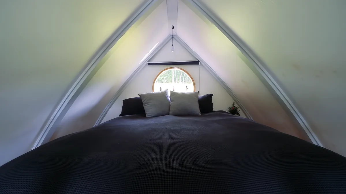 A cozy bed with a dark cover and pillows under a triangular ceiling with a small round window.