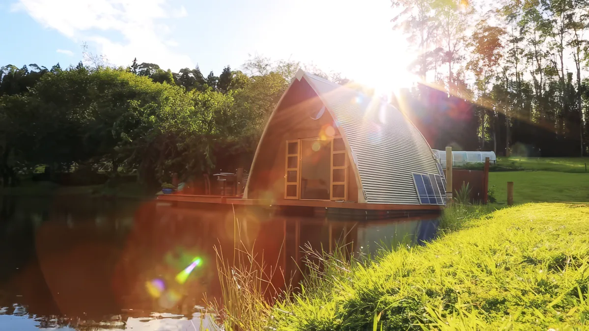 A cozy, dome-shaped cabin sits on a serene lake, surrounded by lush greenery and bathed in golden sunlight.