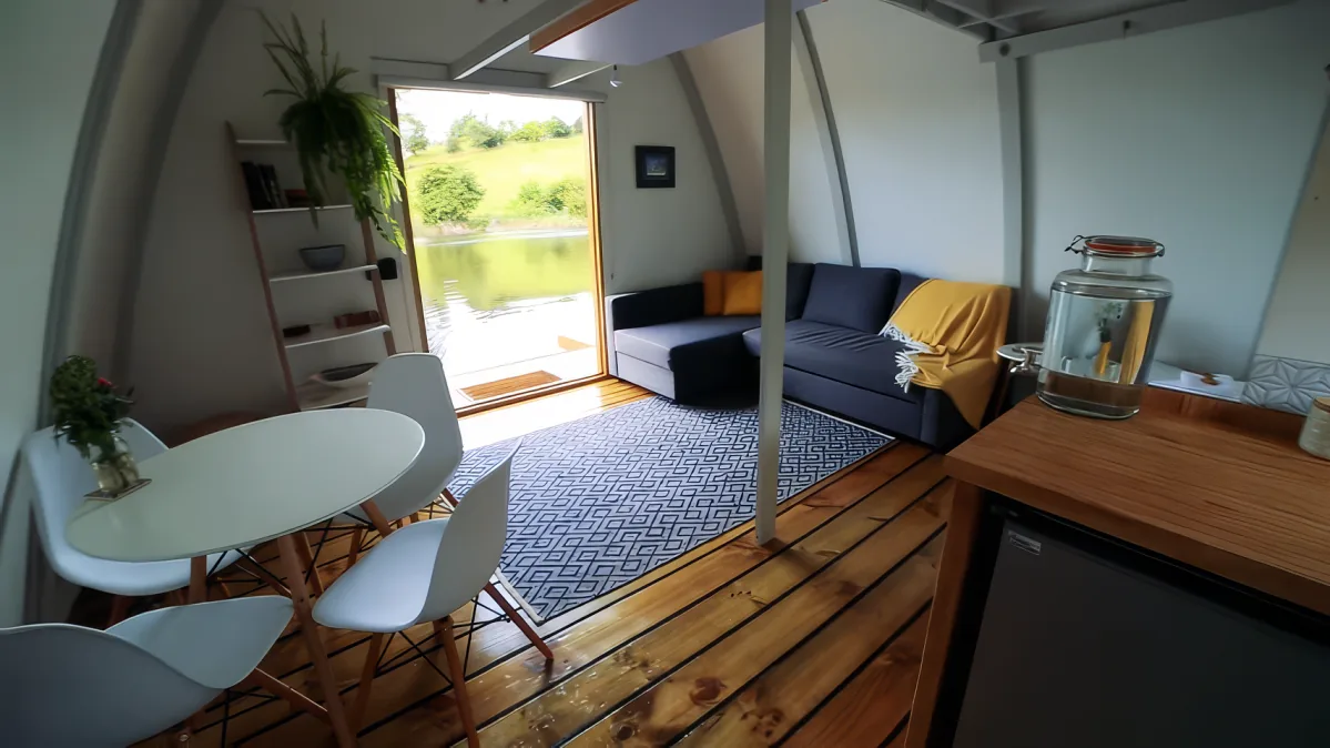 A cozy, modern tiny house with a wooden floor, white chairs, a black sofa, and a view of a serene lake through large windows.