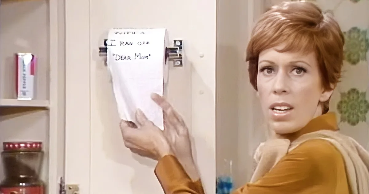 A woman in a yellow outfit holds a note saying "I ran off, Dear Mom" in a kitchen setting.