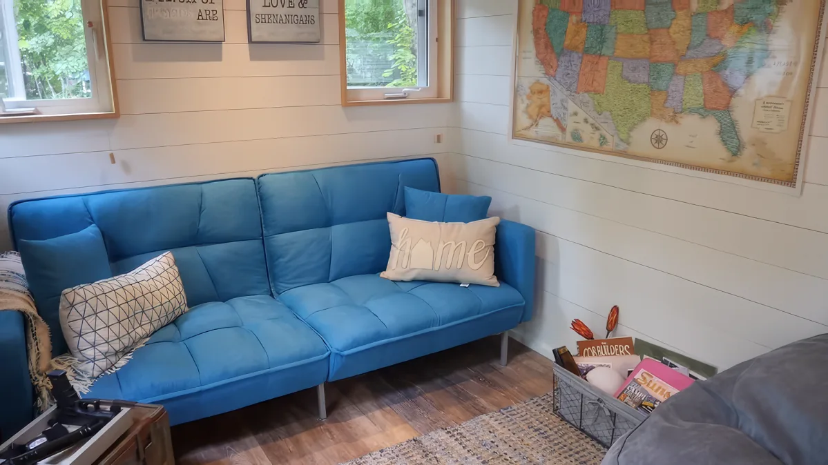 A blue couch with pillows sits in a cozy room with white walls, a map, and a window.