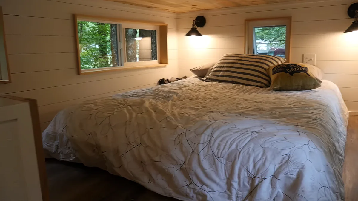 A cozy bedroom with a neatly made bed, pillows, and a quilt, featuring two windows and wall-mounted lamps.