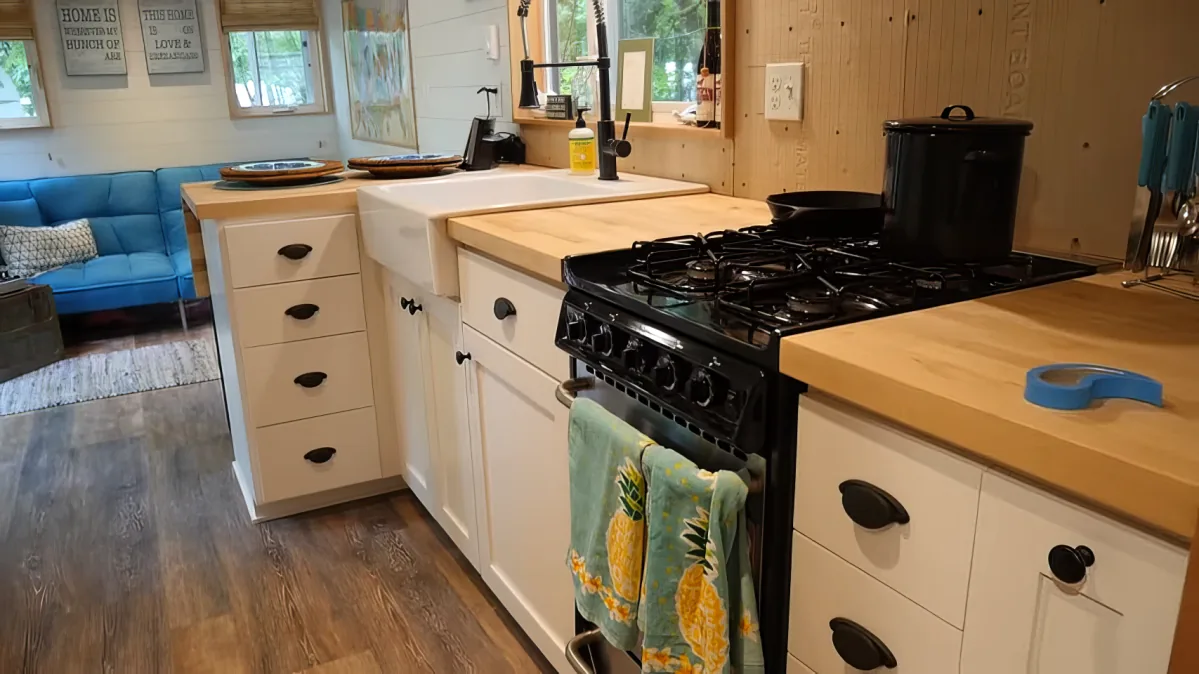 A cozy kitchen with white cabinets, a gas stove, wooden countertops, and a blue couch in the background.