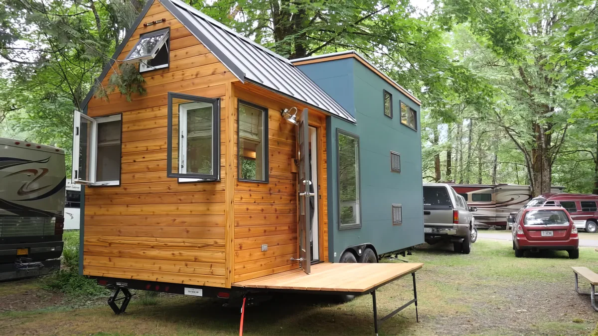 A wooden tiny house with a teal section sits on a trailer amidst a forested area with parked vehicles.