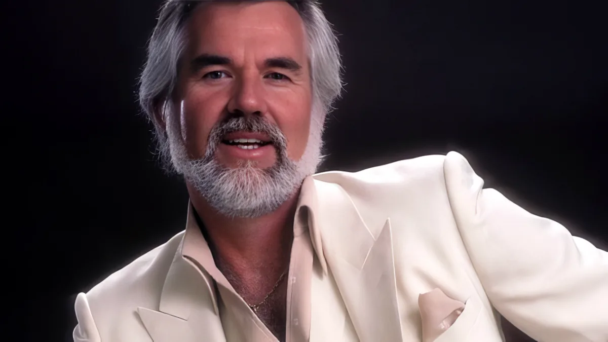 A man with gray hair and a beard, wearing a light-colored suit, is seated against a dark background.