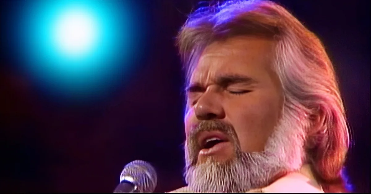 A man with long hair and a beard sings into a microphone under stage lighting.
