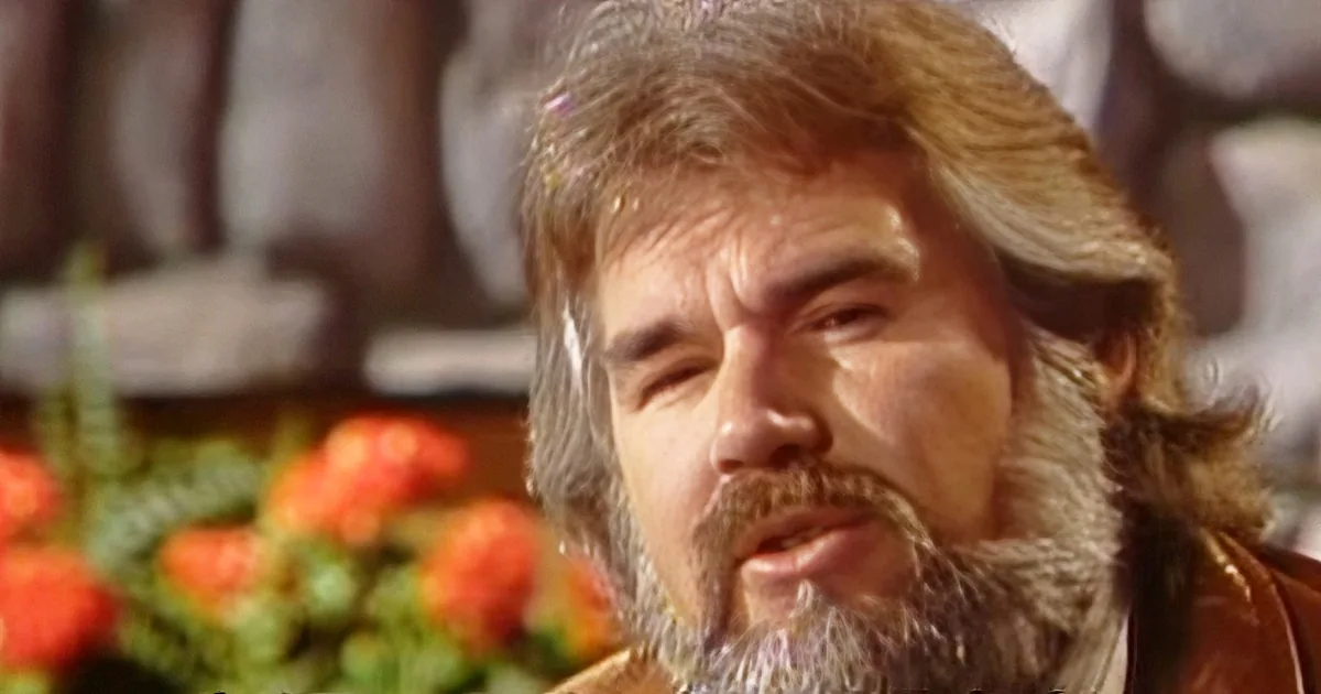 A man with long, wavy hair and a beard, wearing a brown leather jacket, is smiling slightly.