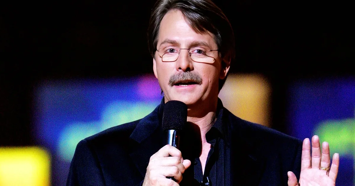 A man in a black suit holds a microphone, gesturing with his hands, against a blurred colorful background.