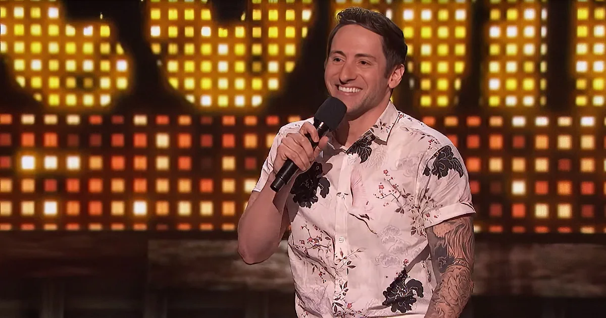 A man in a floral shirt holds a microphone, smiling, with a colorful, illuminated background.