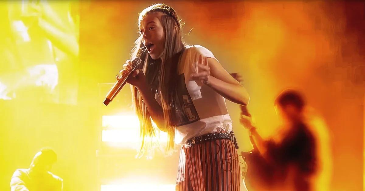 A performer with long hair and a microphone is singing on stage with bright, colorful lighting.