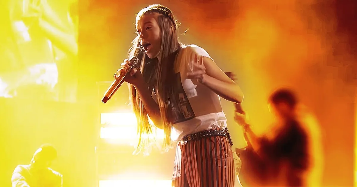 A person with long hair and a white shirt sings passionately into a microphone on a stage with vibrant yellow and orange...
